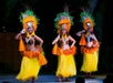 Three women in traditional Hawaiian costumes perform a hula dance on stage, wearing coconut shell tops, yellow grass skirts, and feathered headdresses.