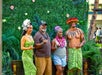 Two people pose with two performers in traditional Polynesian attire in front of a green leafy backdrop decorated with flowers.