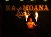 A performer in traditional attire holds two flaming torches in front of a sign that reads "Ka Moana Luau" against a dark background.