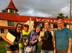 Five people pose outdoors in front of a building with a red roof and the word "KONA" visible; several have surprised or playful expressions and are holding various items.