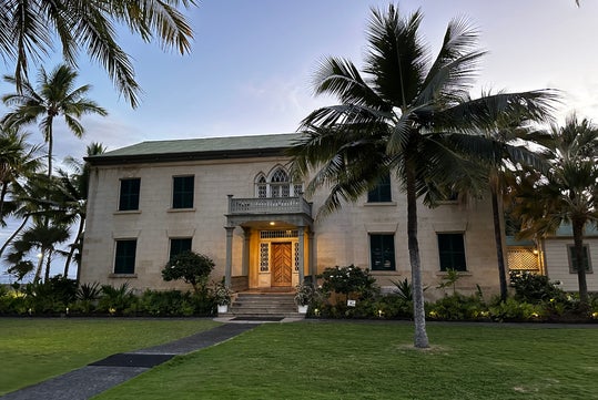 A two-story stone building with green shutters, surrounded by palm trees and lawn, at dusk. The entrance is lit, with steps leading up to a wooden door.