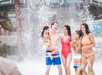 A group of five people in swimsuits stand and smile together in front of a large splash of water at an indoor water park.