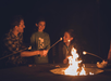 Three people roast marshmallows over an outdoor firepit at night, with two children smiling and holding sticks and an adult assisting.
