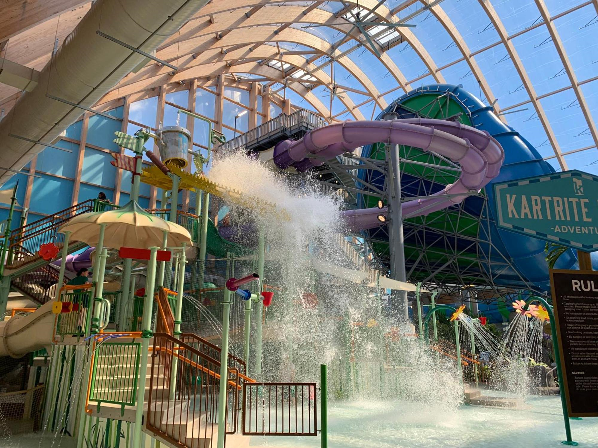 Indoor water park with large purple slides, a water dumping bucket in action, and a bright, sunlit glass ceiling.