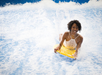 A woman in a white swimsuit rides a yellow bodyboard on artificial waves, smiling as water splashes around her.