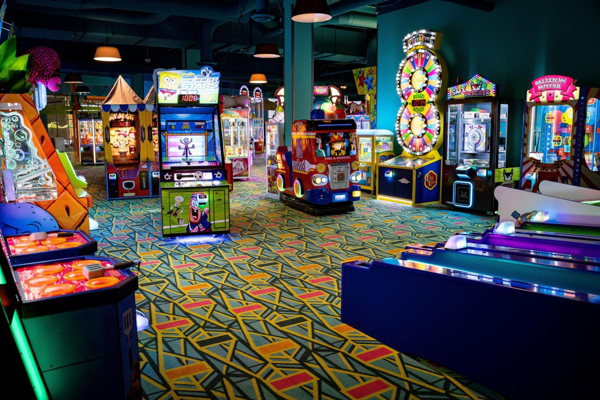 Brightly lit arcade room with colorful patterned carpet, featuring various games and machines, including skee-ball, claw machines, and a kiddie ride shaped like a fire truck.