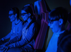 Three people wearing 3D glasses sit in red motion simulator seats, illuminated by blue light, appearing engaged with what they are viewing.