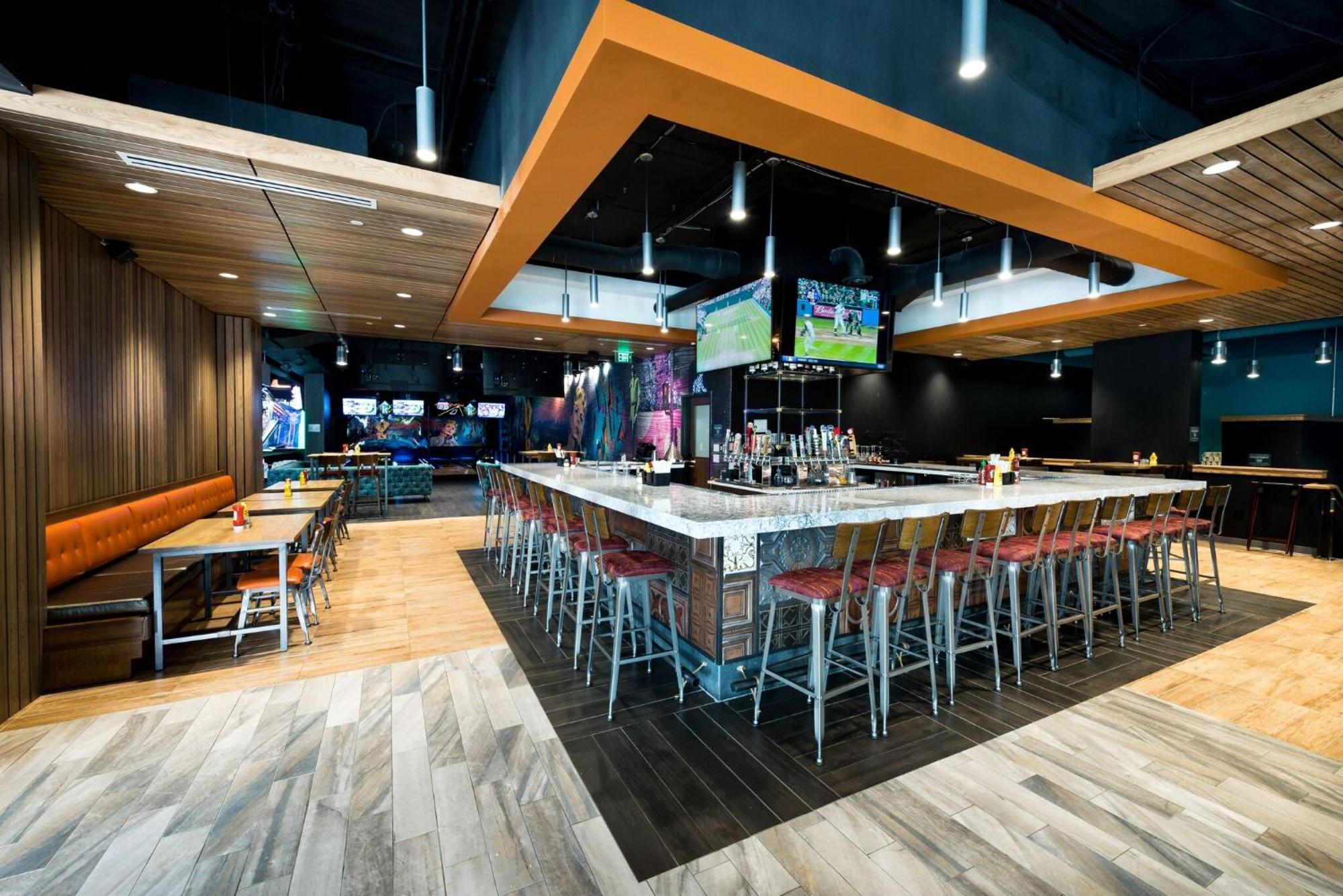 A modern sports bar with a central square bar, high stools, TVs showing sports, and booths along the wall in a well-lit space.