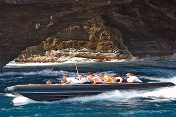 A group of people ride a black inflatable boat through choppy blue water near a rocky sea cave.