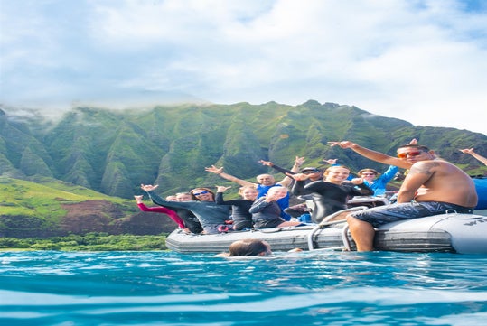 A group of people on an inflatable boat pose and smile in front of green, mountainous cliffs, with blue water in the foreground.