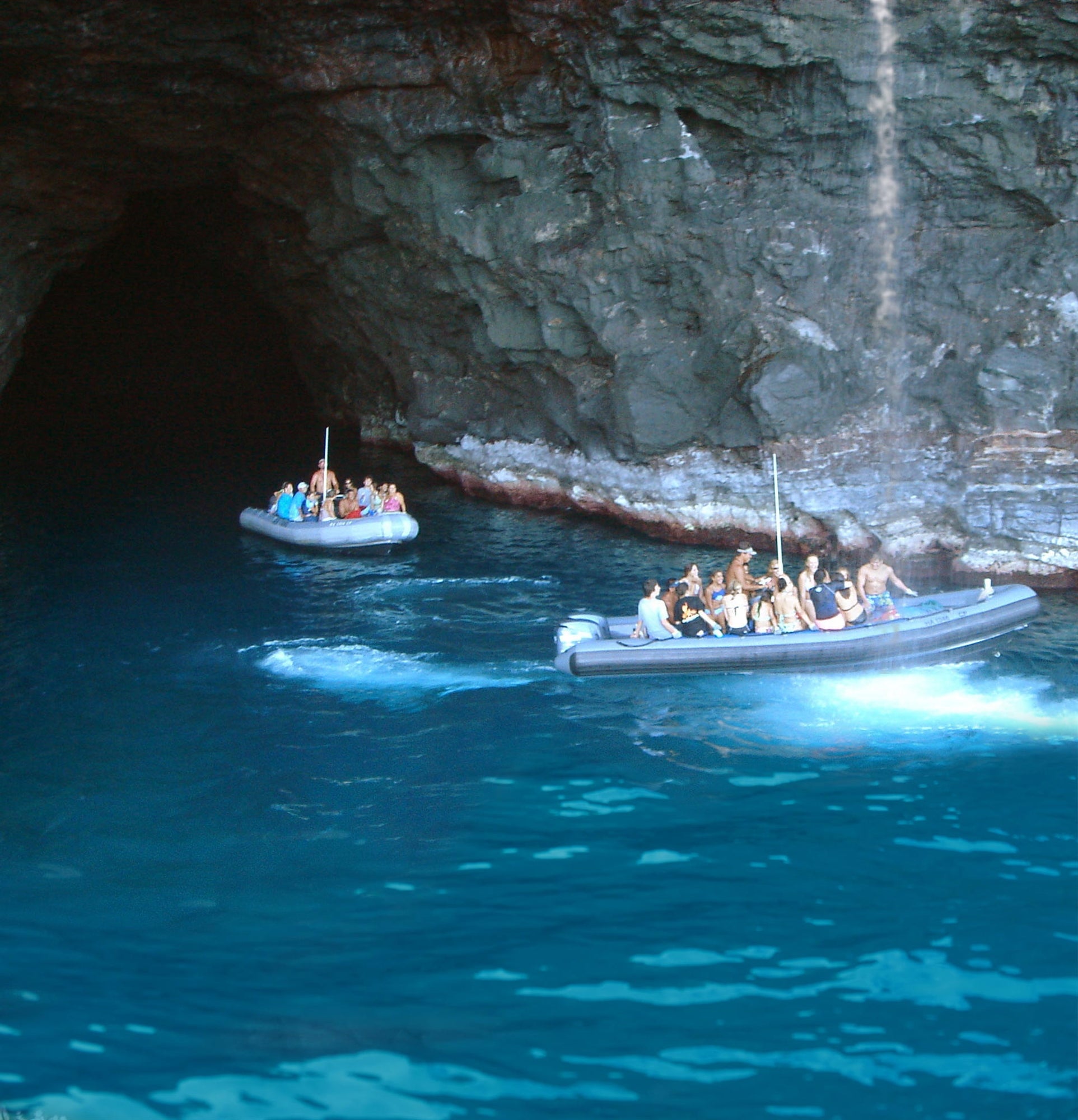 Two inflatable boats with people wearing life jackets approach a large rocky cave opening above deep blue water.