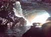 Interior of a rocky cave with sunlight filtering through. Water cascades down the rocks into a pool, and a silhouetted person appears to photograph the scene.