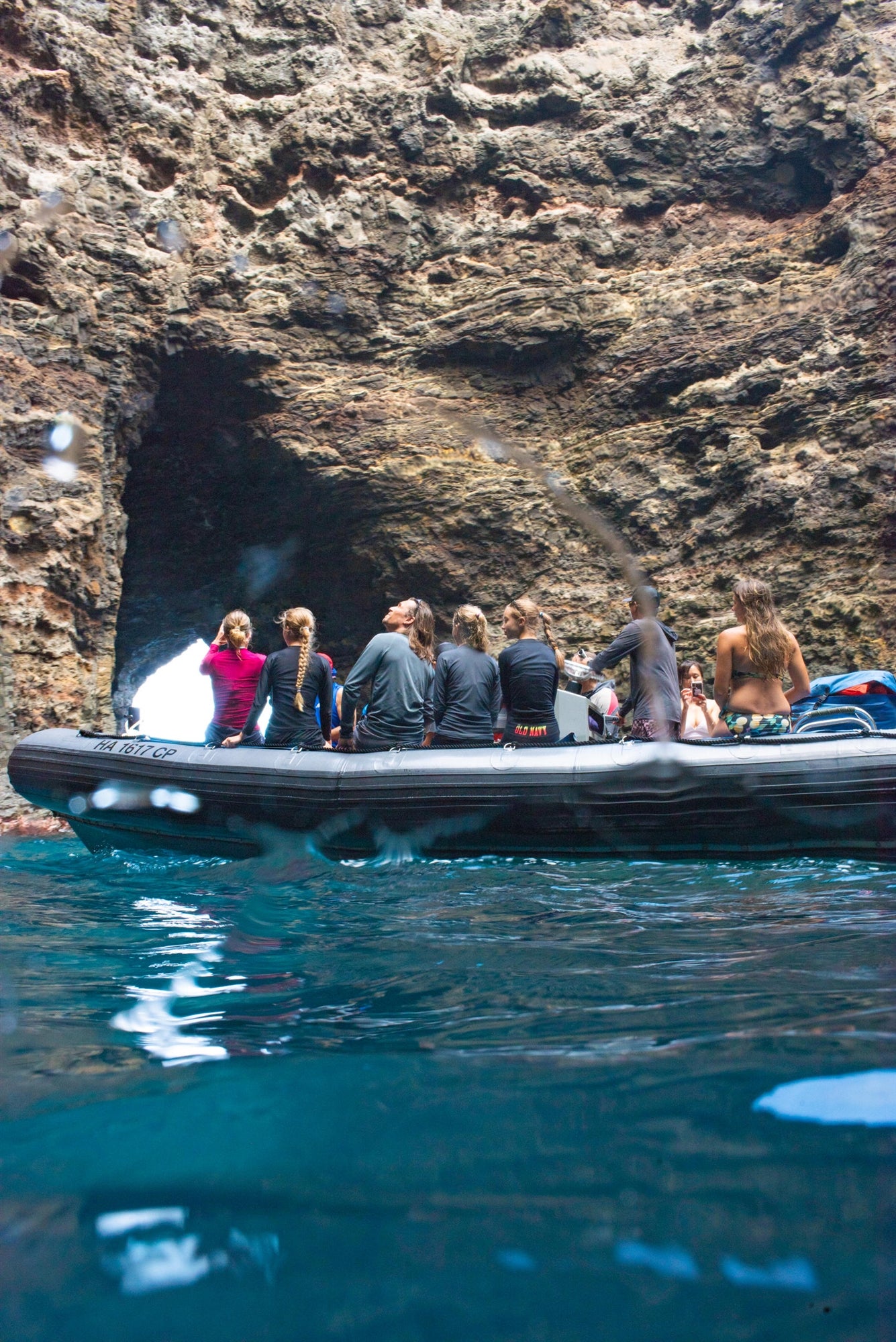 A group of people stand on a boat near a rocky cave entrance, surrounded by water.