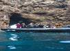 A group of people on an inflatable boat approach a large rocky cave opening along a rugged coastline, with clear blue water in the foreground.