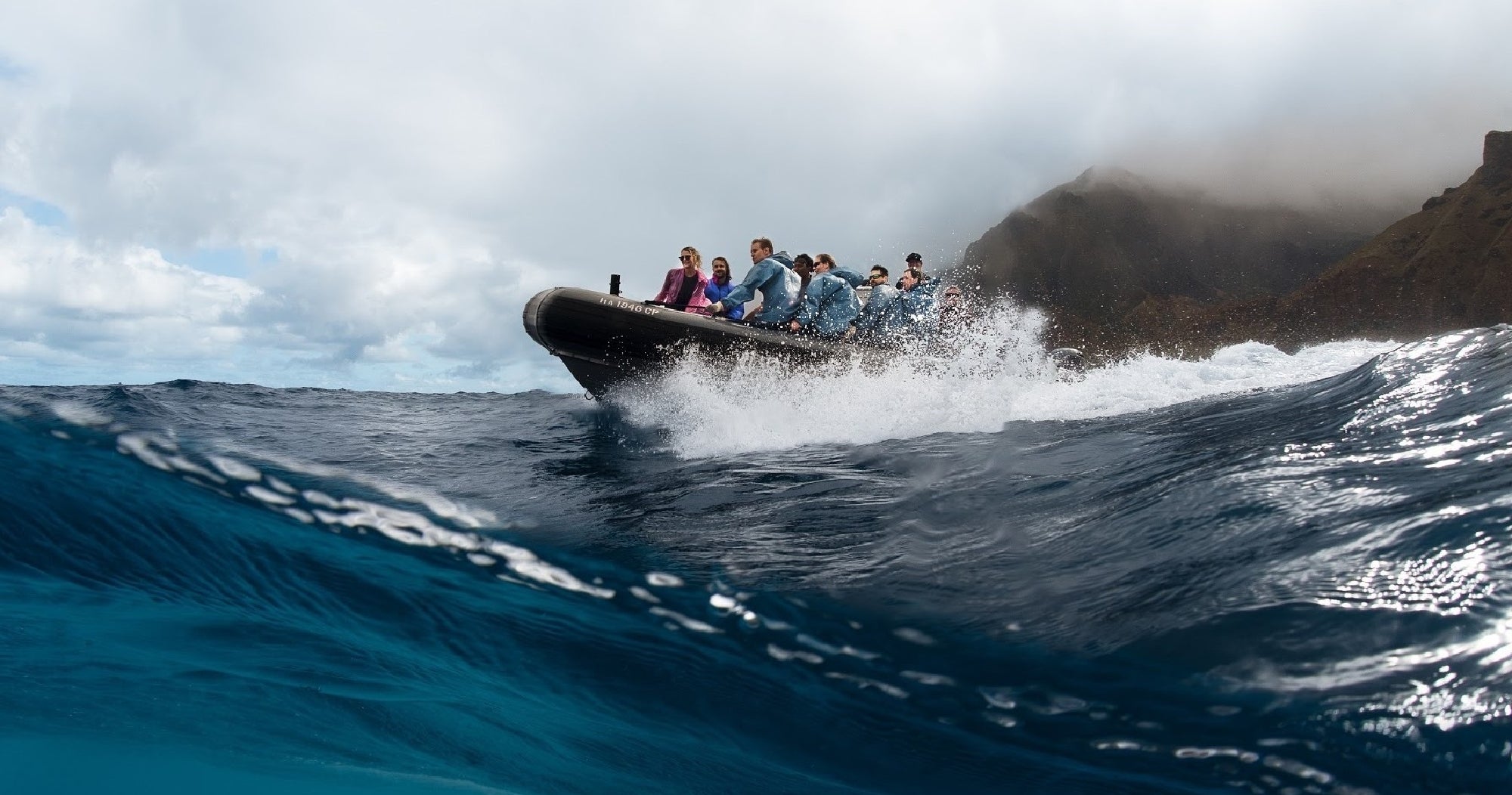 A group of people ride a black inflatable boat over choppy ocean waves near a rocky, fog-covered coastline under a cloudy sky.
