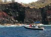 A small inflatable boat with people aboard floats on blue water near a rocky, red and black cliff covered with sparse vegetation.