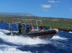 A small motorboat with people on board moves quickly across blue ocean water near a green, hilly shoreline under a partly cloudy sky.