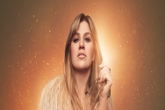 A person with long hair and a fur jacket is illuminated by warm, glowing light against a golden background.