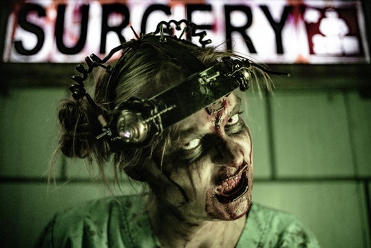 A person in distressed makeup and a head restraint poses under a sign reading "SURGERY," appearing as a zombie or horror character.