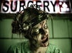 A person in distressed makeup and a head restraint poses under a sign reading "SURGERY," appearing as a zombie or horror character.