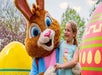 A person in a bunny costume stands beside a smiling young girl. They are surrounded by oversized decorated eggs in an outdoor setting.