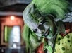 A person dressed as a creepy clown with green hair, white face paint, and a green polka-dot outfit, smiling at the camera in a dimly lit room.