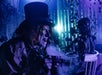 A person in a top hat and creepy makeup holds a smoking cup in a dimly lit, eerie setting with blue and purple lighting.