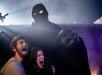 Two people scream in fear with a hooded figure in dark robes and glowing eyes looming behind them in a foggy, dimly lit setting.