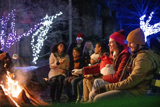 A group of people sit around a campfire at night, roasting marshmallows, with trees decorated with string lights in the background.