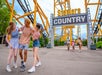 Three friends laugh and walk under the "Steelers Country" sign at an amusement park with yellow roller coaster tracks in the background.