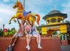 Three people sit on a ledge taking a selfie outside the entrance of Kennywood amusement park, with a large, colorful carousel horse in the background.
