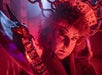 Person in dramatic makeup and costume with playing cards in hair and holding a large staff, illuminated by red lighting.