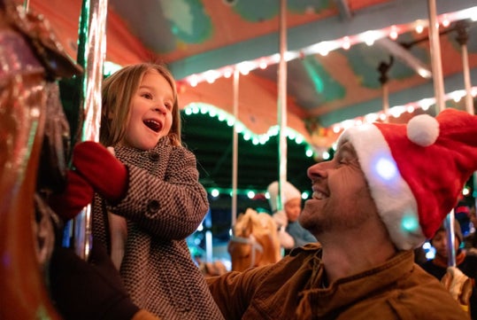 A child and an adult wearing a Santa hat enjoy a carousel ride, surrounded by festive lights and decorations.