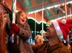 A smiling child in a coat and red mittens rides a carousel next to an adult wearing a Santa hat, both appearing joyful under festive lights.