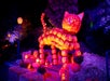 A large cat sculpture made of glowing carved pumpkins stands on hay bales, surrounded by smaller pumpkins, at night.