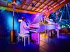 Two people play white grand pianos on a stage under a roof, surrounded by colorful lights and festive decorations.