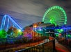 Night scene of an amusement park with a glowing green Ferris wheel, a blue-lit roller coaster, trees, and empty gated walkways in the foreground.