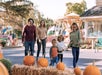 A family of five walks together outdoors near pumpkins and hay bales, with a carousel and trees in the background on a sunny day.