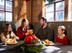 A family of four sits at a festive table with food and drinks, smiling and toasting together in a sunlit, decorated wooden room.