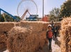 Children walk through a hay bale maze at an outdoor fairground with a Ferris wheel and trees visible in the background on a sunny day.