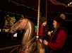 A young child sits on a carousel horse, smiling at an adult beside them. Both wear warm clothing, and festive lights glow in the background.