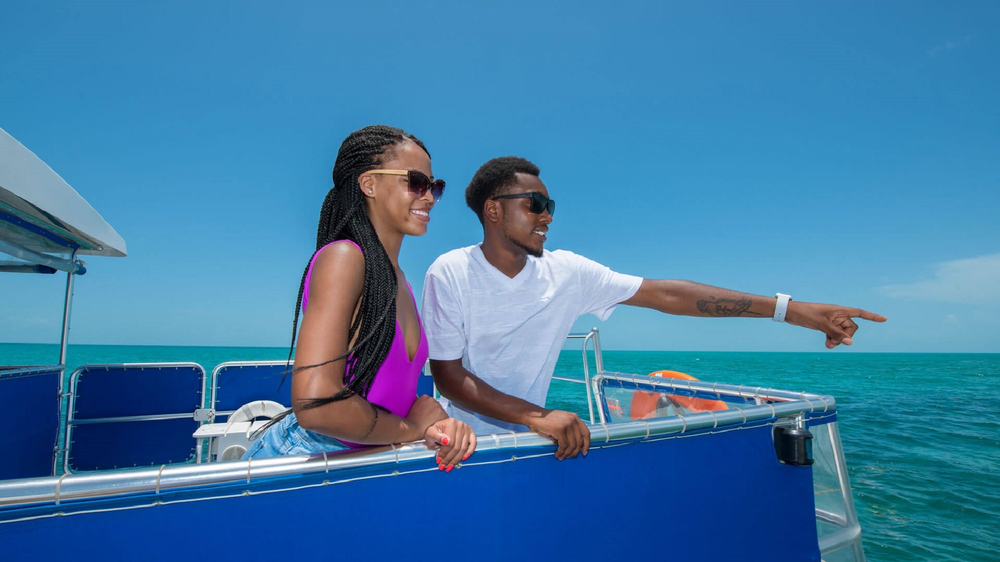 Two people stand on the deck of a blue boat, enjoying the view of the ocean. The man points towards the horizon while the woman smiles beside him under a clear, sunny sky.