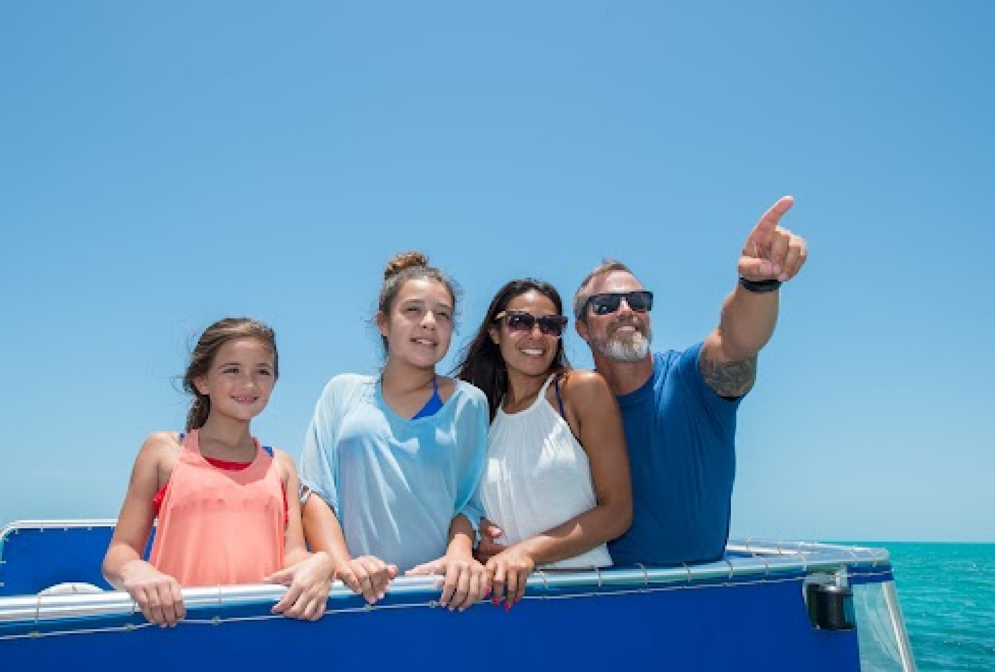 A family of four, including two children and two adults, is on a boat in sunny weather. One adult is pointing towards the horizon. They are all smiling and enjoying the sea.