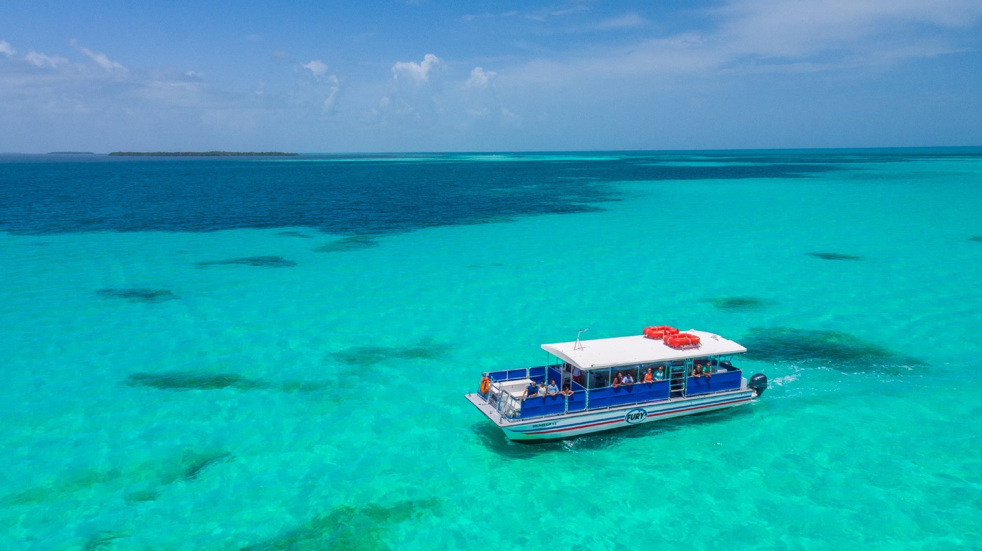 A small boat with people onboard is cruising through vibrant turquoise waters under a clear blue sky. An island is visible in the distance.