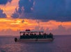 A boat with passengers sails on calm waters at sunset, under a vibrant sky with orange and purple clouds.