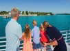 Four people, two adults and two children, stand on a boat looking at the shoreline across turquoise water on a sunny day. One child points toward the land.