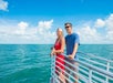 A woman in a red dress and a man in a blue shirt stand on a boat deck overlooking the ocean under a blue sky with scattered clouds.