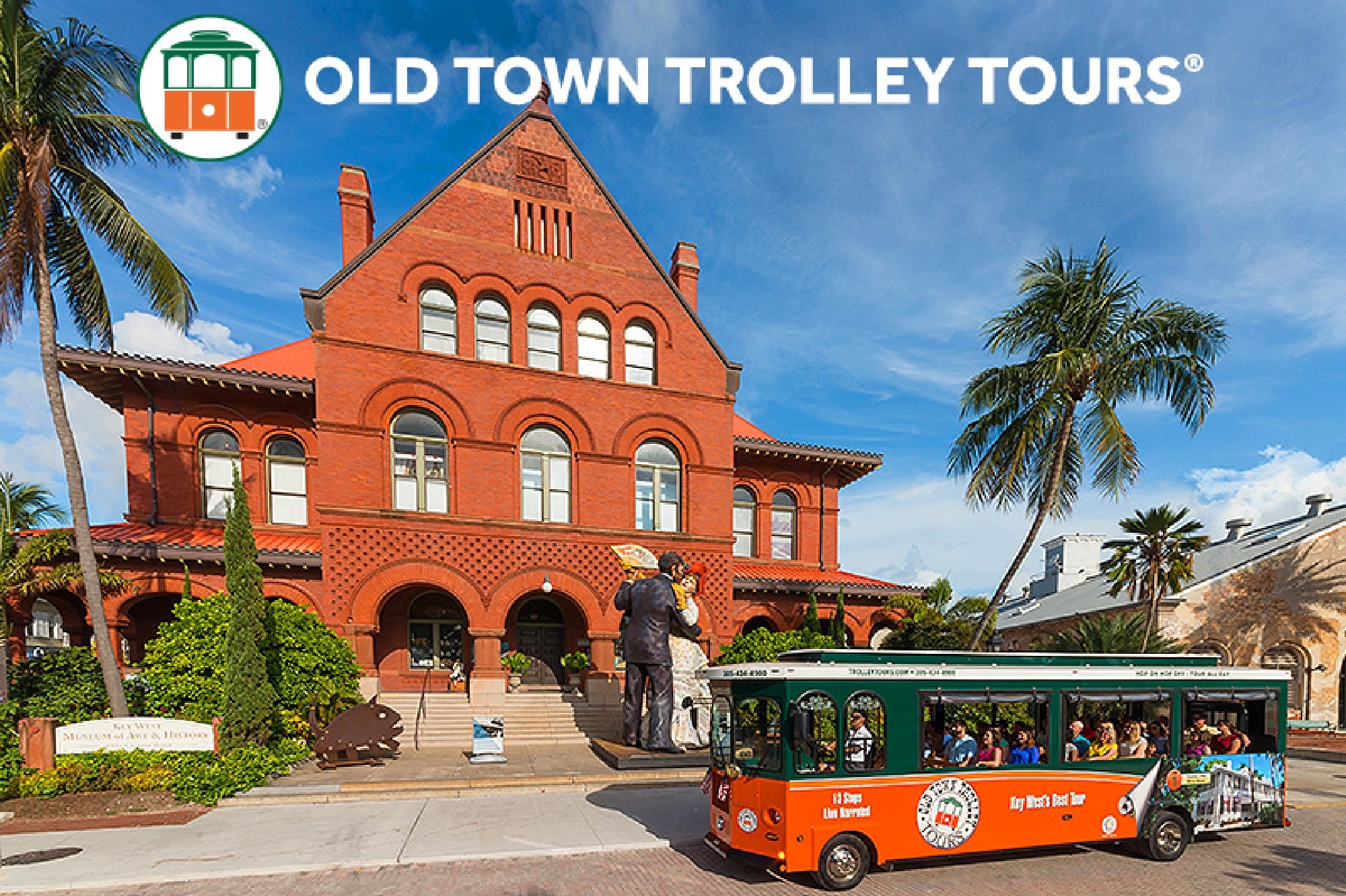 A green Old Town Trolley bus parked in front of a large red brick building with palm trees under a blue sky.