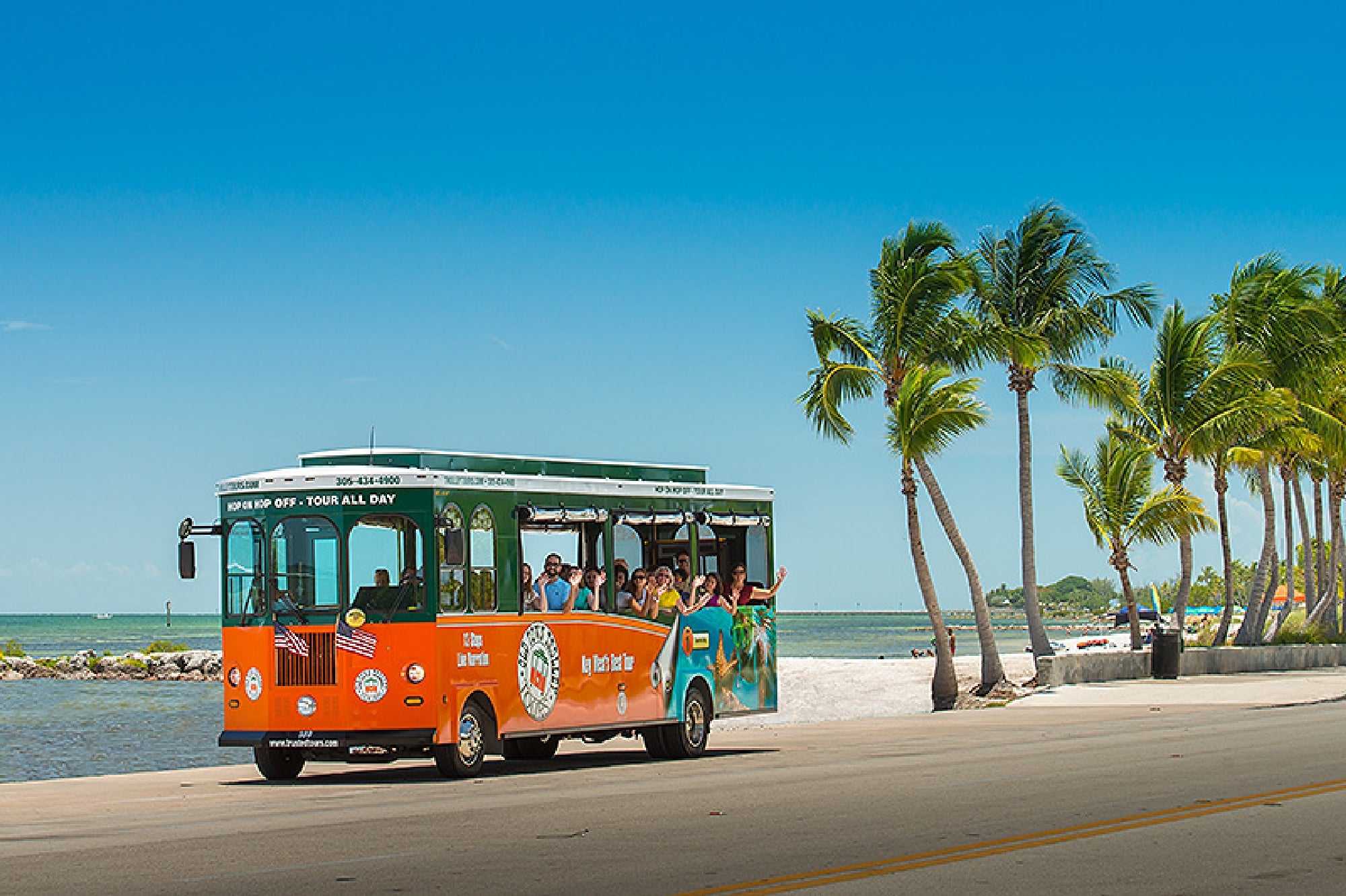 An orange and green open-air trolley with passengers drives along a palm-lined road by the ocean under a clear blue sky.