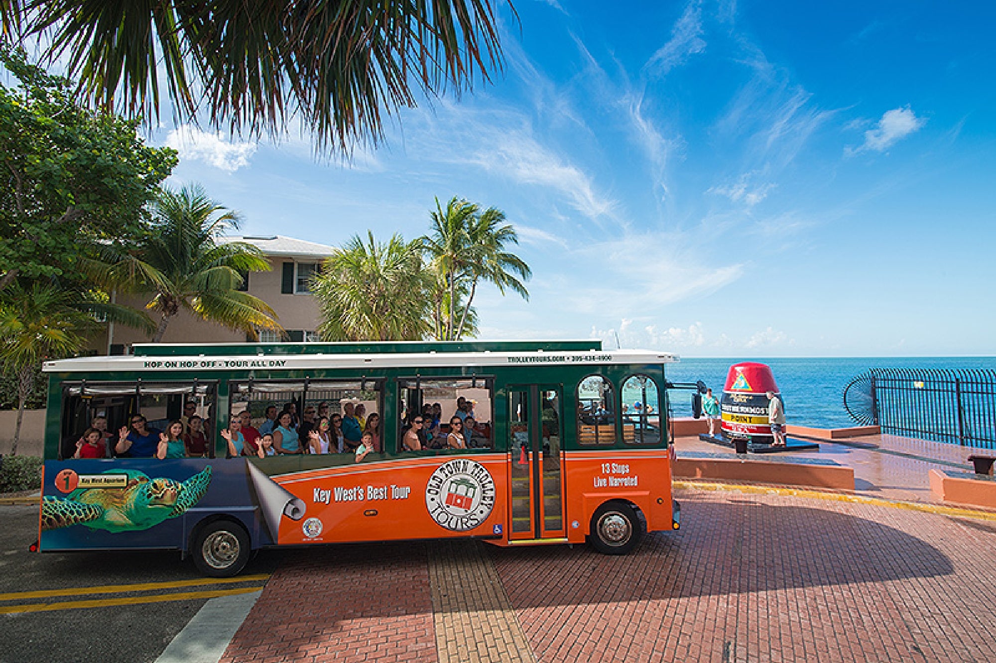 A red and green sightseeing trolley with tourists drives past the Southernmost Point Buoy in Key West, Florida, near the ocean on a sunny day.
